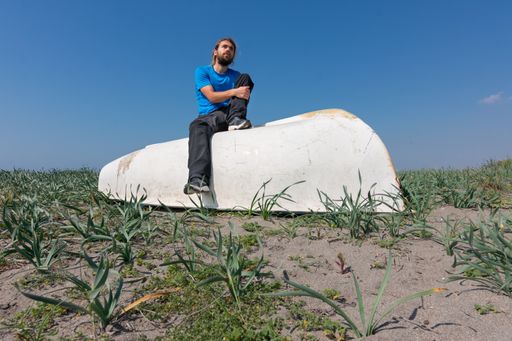 A bearded caucasian male in a blue shirt sitting on a white painted big object with a clear blue sky in the background