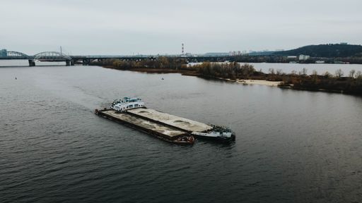 An aerial view of cargo ship sailing along the Dnieper river with Darnitsky bridge in the distance in Ukraine, Kyiv