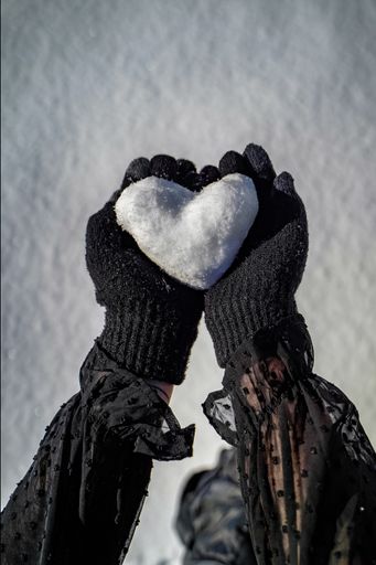 A vertical top view of a person's hand in black gloves holding a heart made of snow