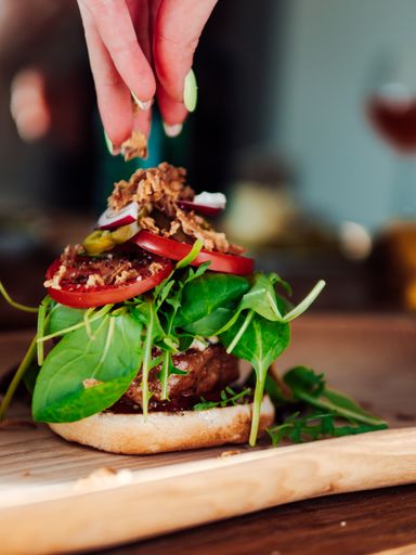 A female hand dressing a yummy burger with meat, salad and other ingredients on a wooden surface