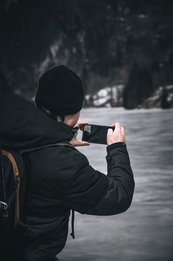 Vertical shot of a person from behind wearing black jacket and a beanie and taking photos with his mobile