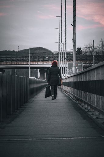 A vertical shot of a woman from behind wearing a black jacket and walking on a bridge in a city