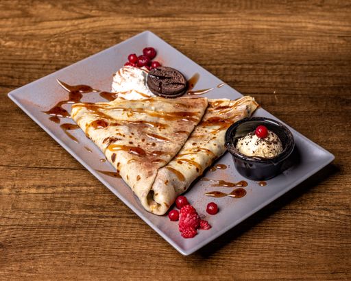 A plate of crepe with caramel sauce drizzles, whipped cream and a biscuit on a wooden table