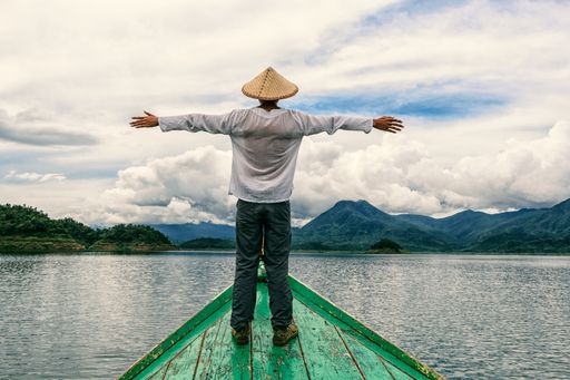A man wearing a chinese hat standing with open hands on the edge of a boat in the Myo Gyi Dam, Myanmar
