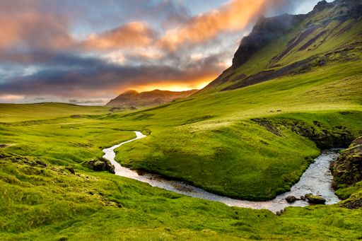 A high angle shot of a river in the middle of grassy hills near a mountain under a cloudy sky