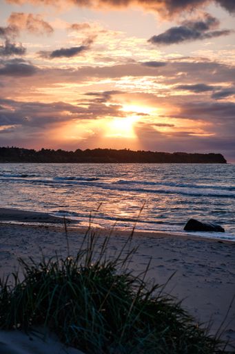 A vertical shot of the ocean with the golden sun setting on the horizon with clouds covering it