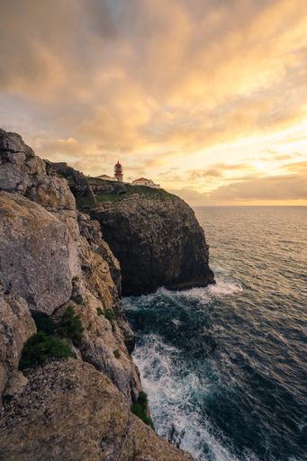 A vertical shot of a lighthouse on rocky cliffs by the sea outside Sagres in Algarve, Portugal during sunset