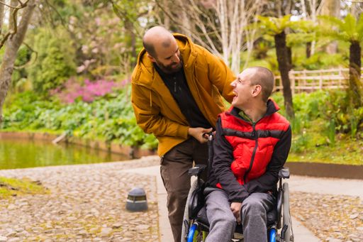 A paralyzed young male in a wheelchair being pushed by a friend in a sunny park