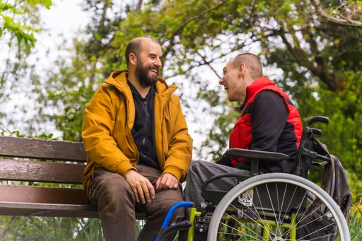 A paralyzed young male in a wheelchair and his friend having fun on a bench in a park