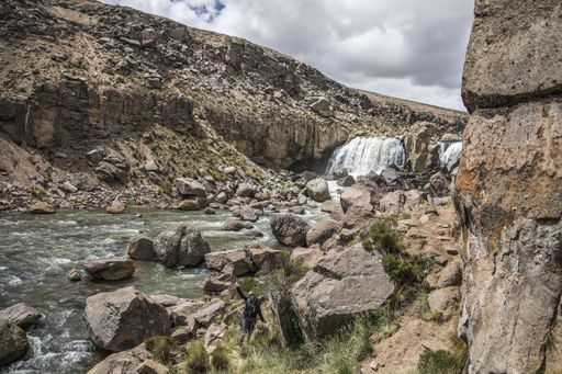 A mesmerizing shot of a tourist standing in front of a  wild waterfall under the cloudy vibrant sky