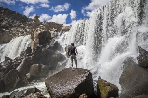 A mesmerizing shot of a tourist standing in front of a  wild waterfall under the cloudy vibrant sky