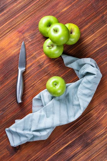 A high angle shot of some green apples and a knife and a tablecloth on a wooden surface