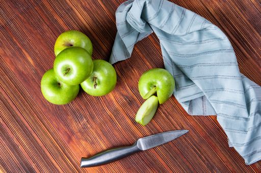 A high angle shot of some green apples and a knife and a tablecloth on a wooden surface
