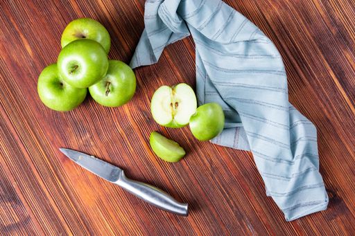 A high angle shot of some green apples and a knife and a tablecloth on a wooden surface