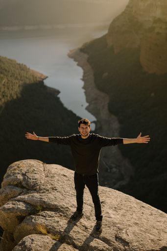 A vertical shot of a young Hispanic man standing on a huge rock with his arms open.