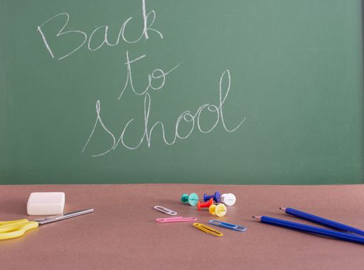 The phrase "back to school" written on a green chalkboard behind the desk with pencils and pins