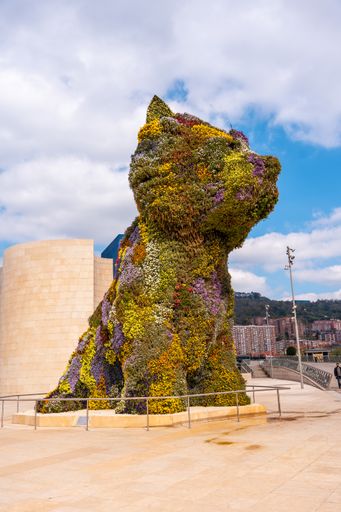 A vertical shot of the colorful Puppy sculpture by Jeff Koons outside the museum Guggenheim museum, with tourists photographing beside it, Bilbao