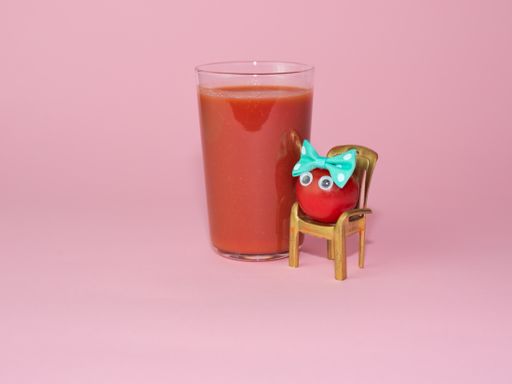 A cup of tomato juice and a whole cherry tomato with eyes on a small chair isolated on pink background