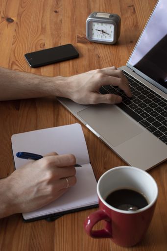 A vertical shot of a male hand using his laptop with a clock, phone, notebook and a cup of coffee on a wooden table
