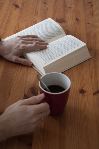 A vertical shot of a male hand holding a cup of coffee while reading a book on a wooden table