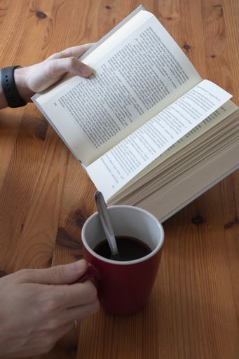 A vertical shot of  a male hand holding a cup of coffee while reading a book on a wooden table