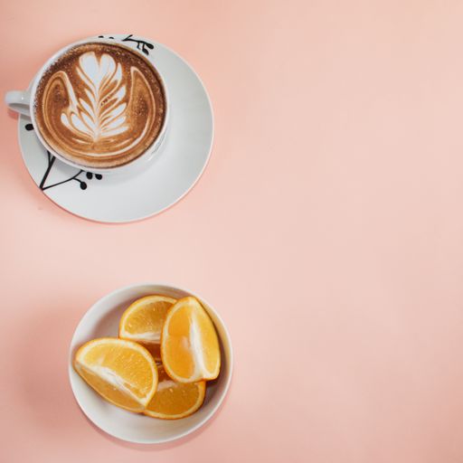 A high angle shot of a cup of cappuccino and sliced oranges in a white bowl against a pink background