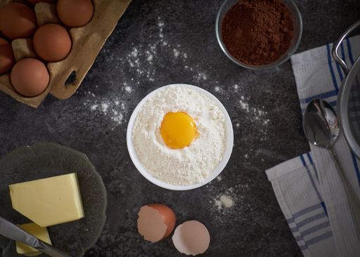 A top view of eggs and flour in a bowl, butter and chocolate powder on a table