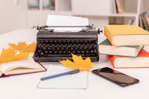 There is retro typewriter on white table. Nearby are books, notebook with pen, mobile phone and yellow maple leaves. Workplace of writer.