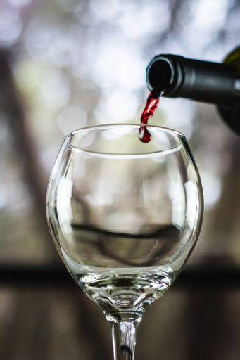 Red wine being poured from a bottle into a wine glass with a stem in front of an open window