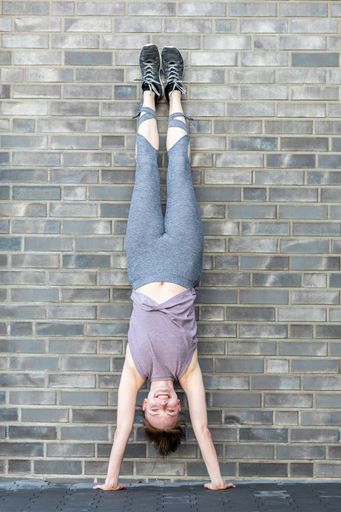 A young Caucasian woman doing a handstand in front of a stone wall as a fitness workout