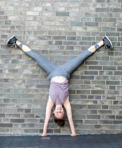 A young Caucasian woman doing a handstand in front of a stone wall as a fitness workout