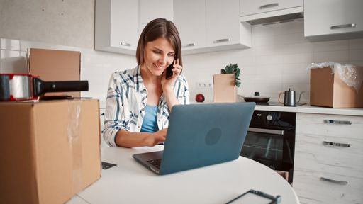 Caucasian young woman working on her laptop in the kitchen at home with boxes on the table and the counter