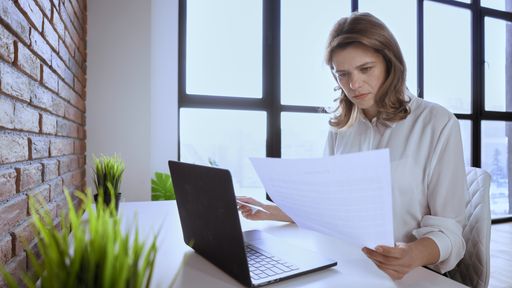 Caucasian young woman sitting at the table and working on her laptop with a paper on her hand