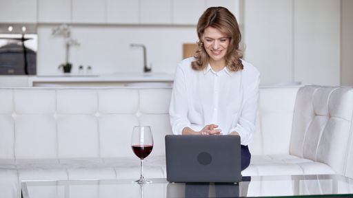 Caucasian young woman sitting on a white coach at home working on her laptop with a glass of wine on the table