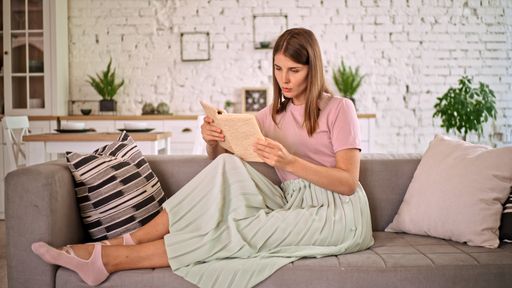 A Caucasian female in a pink t-shirt reading a book sitting on a sofa