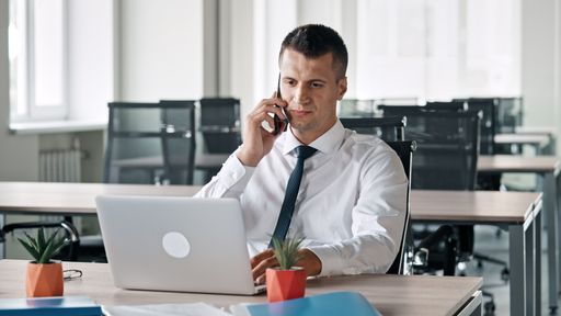 A Caucasian business man sitting in his office and talking on phone with laptop in front of him