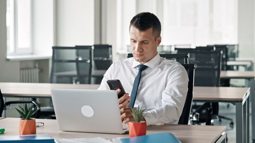 A Caucasian business man sitting in his office and using his phone with laptop in front of him
