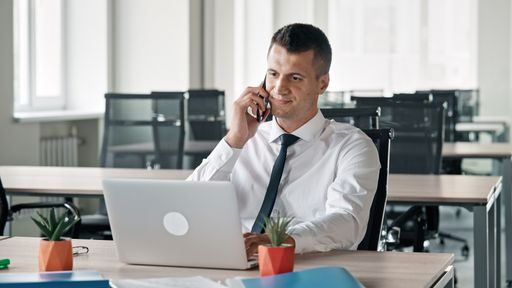 A Caucasian business man sitting in his office and talking on phone with laptop in front of him