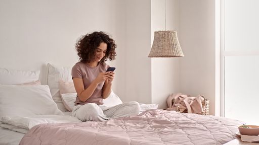 A beautiful shot of Caucasian girl with curly hair smiling and using her phone while sitting on bed