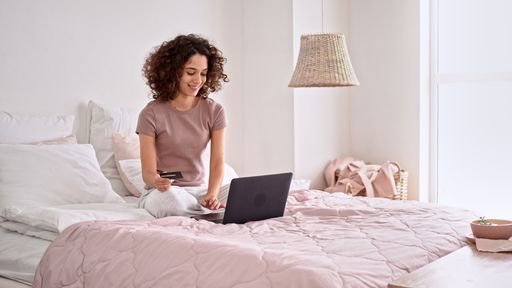 A beautiful shot of Caucasian girl with curly hair smiling and using her laptop while sitting on bed