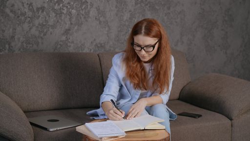 A beautiful Caucasian girl sitting on sofa and writing with laptop and phone next to her