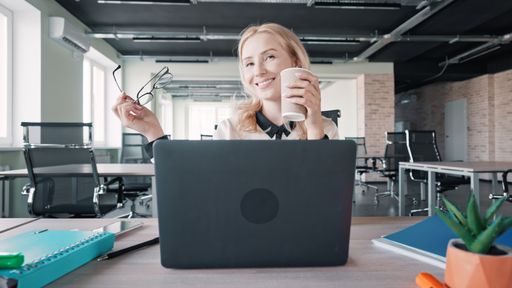 A closeup shot of Caucasian woman sitting on her office and laptop in front of her, holding cup and glasses with smiling