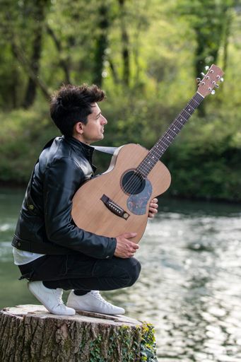 A vertical shot of a caucasian man with a guitar kneeing on a wooden stump by the lake