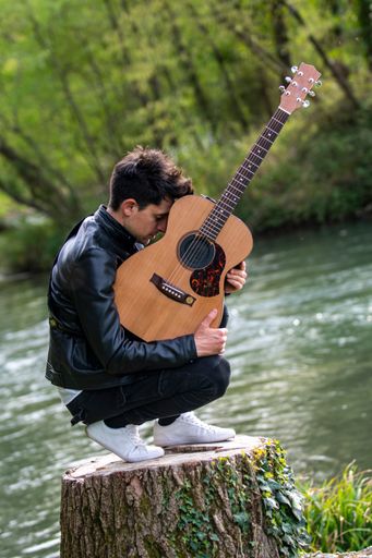 A vertical shot of a caucasian man with a guitar kneeing on a wooden stump by the lake