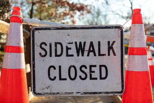 A sign with text "Sidewalk closed" between two traffic cones in the street