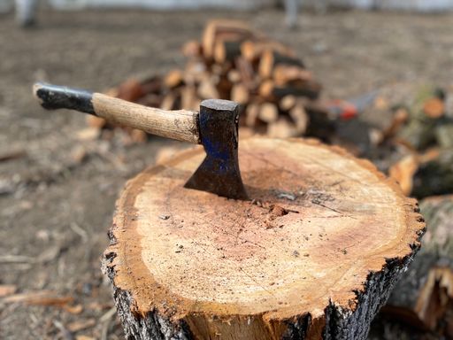 A small ax in a wood log with a pile of lumber in the back.