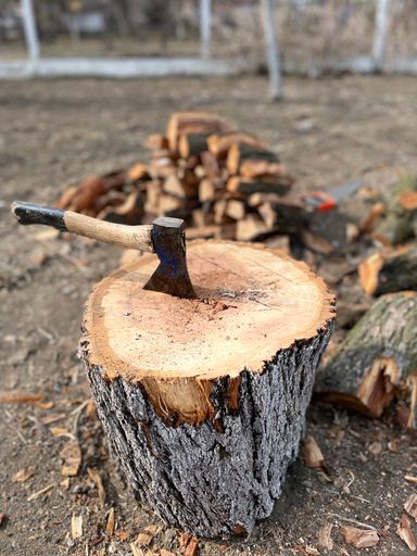 A vertical shot of an ax in a wood stump with a pile of lumber in the background.