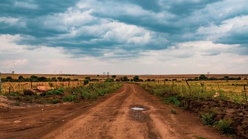 An empty sand road surrounded by fields under a cloudy sky