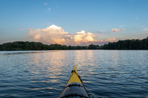The front part of the kayak floating on the wave water surrounded by dense trees at sunset