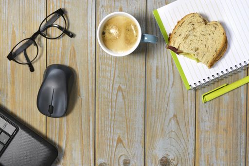 The wireless mouse, optical eyeglasses, cup of coffee, piece of bread and notebook with pen isolated on a wooden surface - work at home concept
